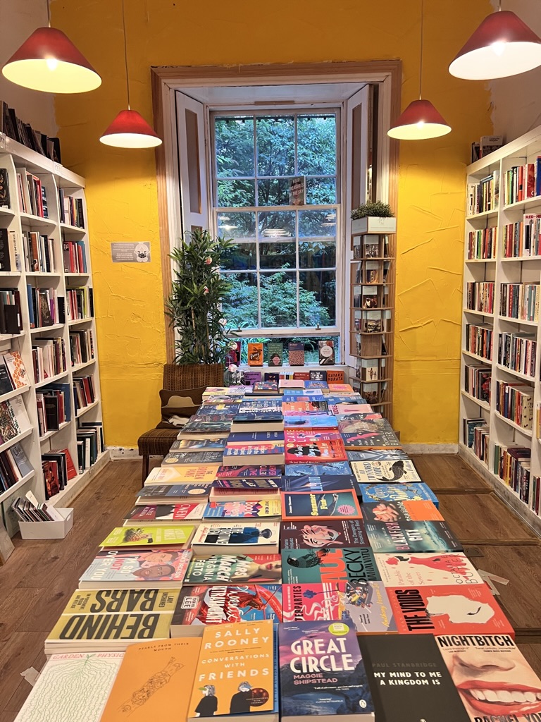 Interior of a bookstore with shelves, a long table of books, a plant and a window looking out to leafy trees